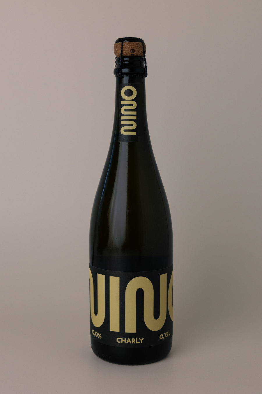 A dark glass bottle of Nino Charly sparkling beverage with a cork stopper, featuring a black label with large gold lettering, set against a solid warm gray background.