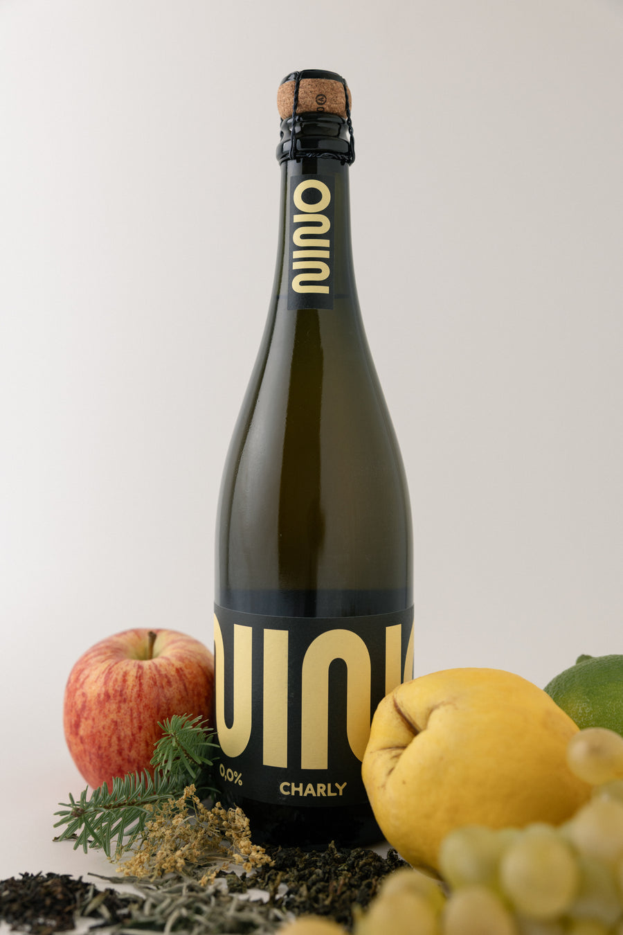 A dark green glass bottle of Nino Charly sparkling beverage with a black label and gold lettering, styled with an apple, lemon, grapes, and dried botanicals on a light gray background.