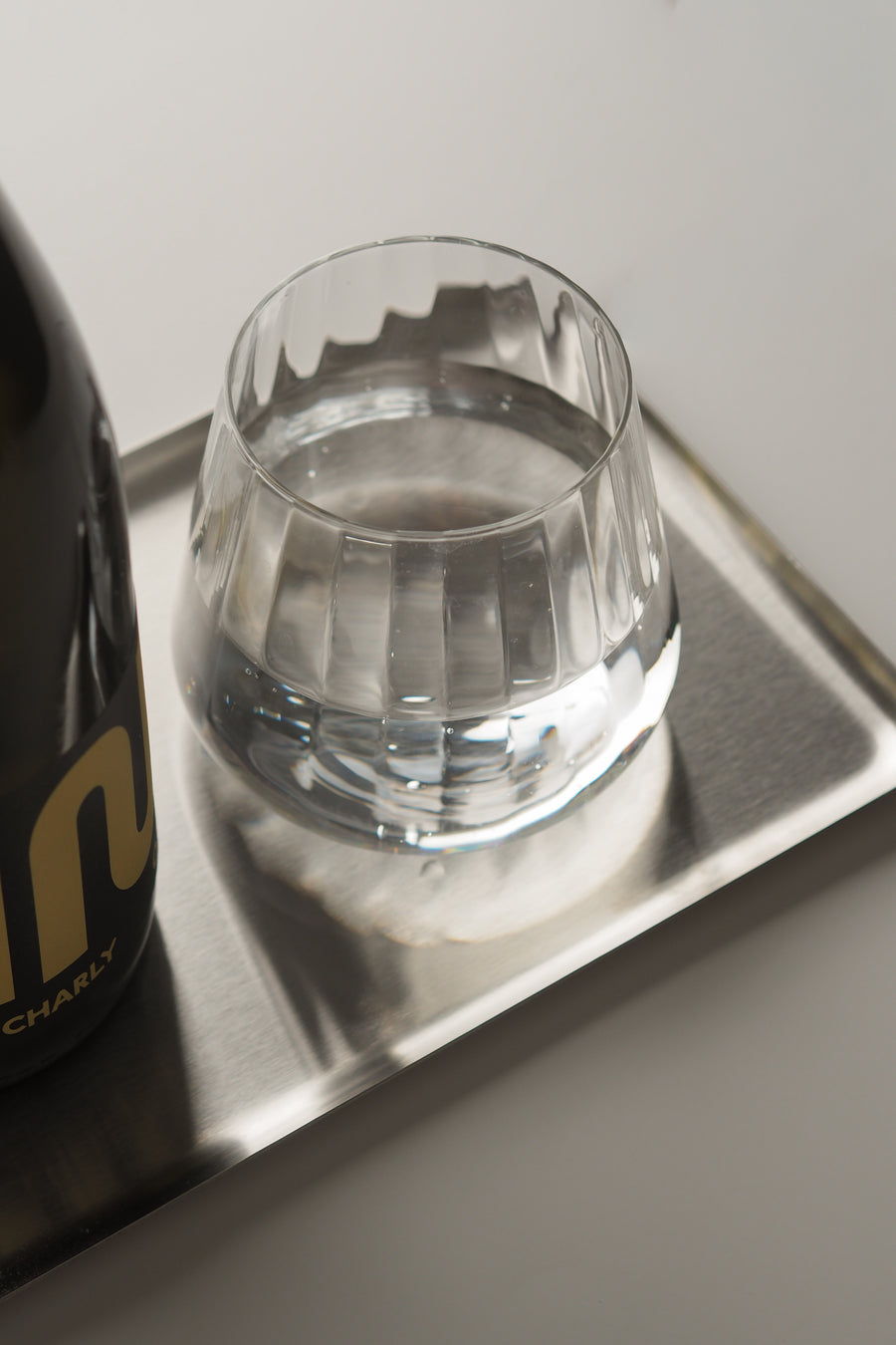 A clear faceted glass tumbler containing water, placed on a silver rectangular tray next to a dark bottle with gold lettering, against a light gray background.