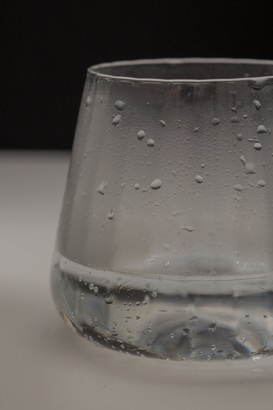 A close-up of a clear glass tumbler with condensation droplets on the outside, set against a dark background and light surface.