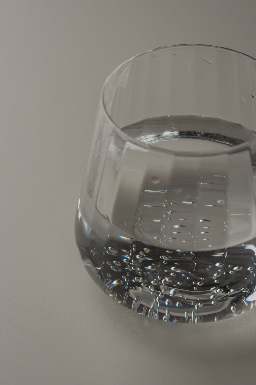 A close-up of a clear glass tumbler containing sparkling water, photographed on a plain light gray background.