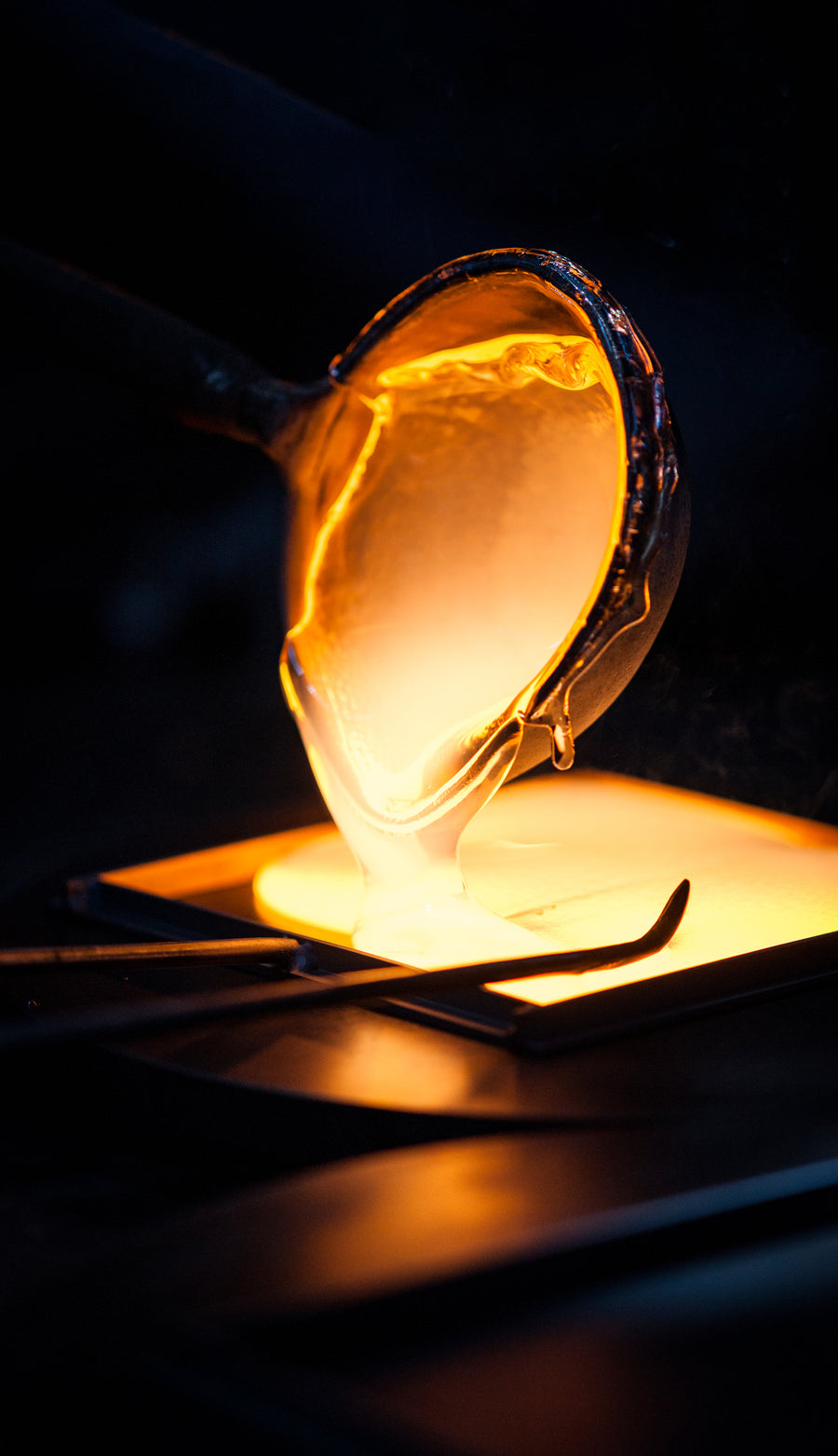 Molten metal being poured from a glowing crucet into a casting mold, shown in dramatic close-up against a dark background.