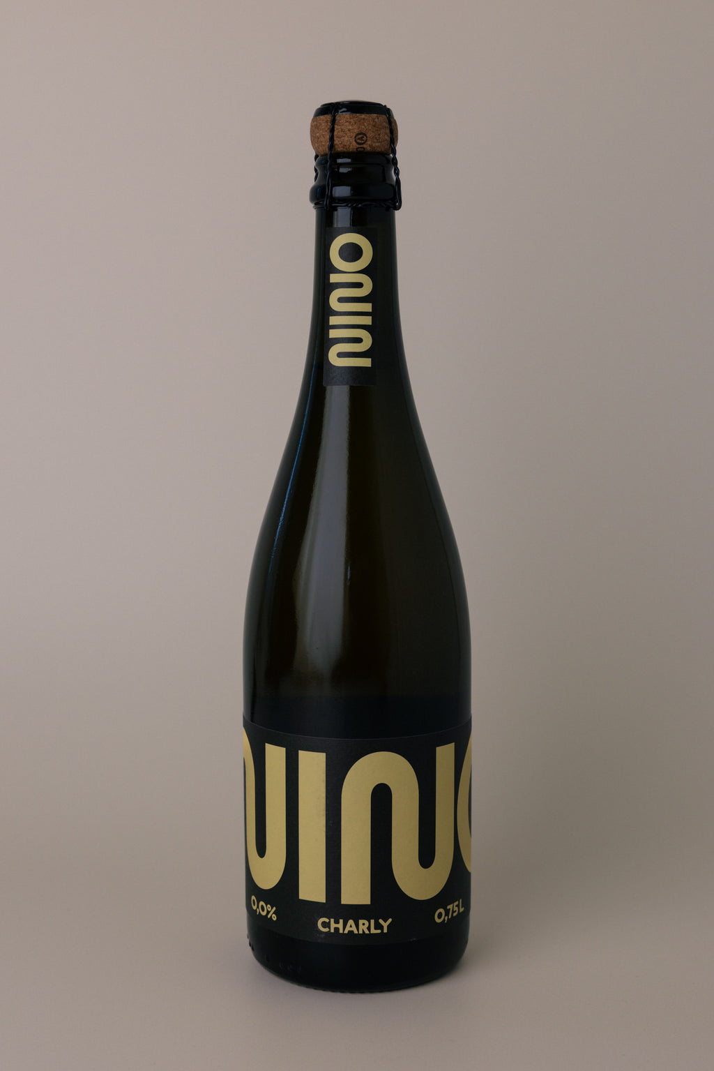 A dark glass bottle of Nino Charly sparkling beverage with a cork stopper, featuring a black label with large gold lettering, set against a solid warm gray background.