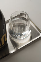 A clear faceted glass tumbler containing water, placed on a silver rectangular tray next to a dark bottle with gold lettering, against a light gray background.