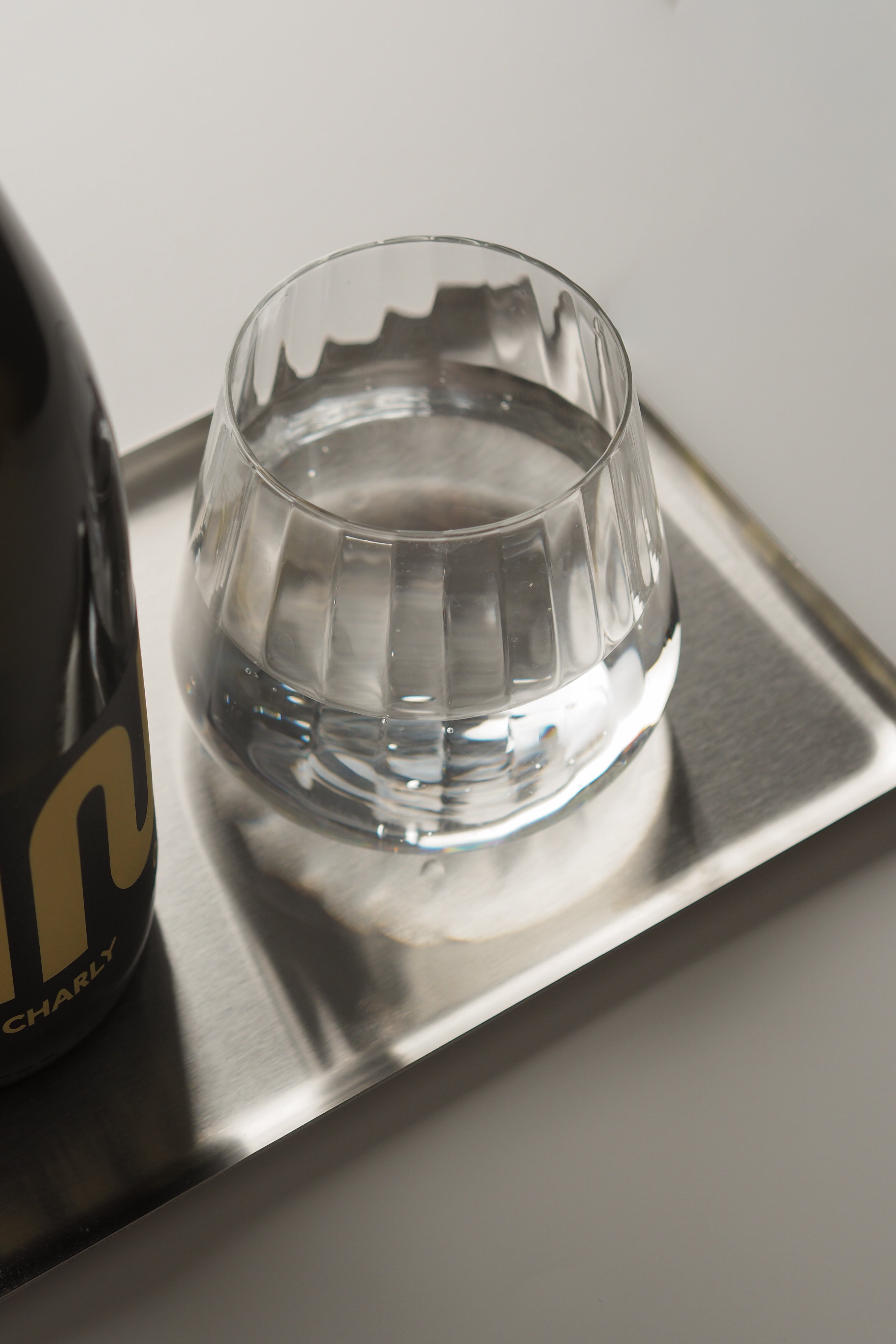 A clear faceted glass tumbler containing water, placed on a silver rectangular tray next to a dark bottle with gold lettering, against a light gray background.