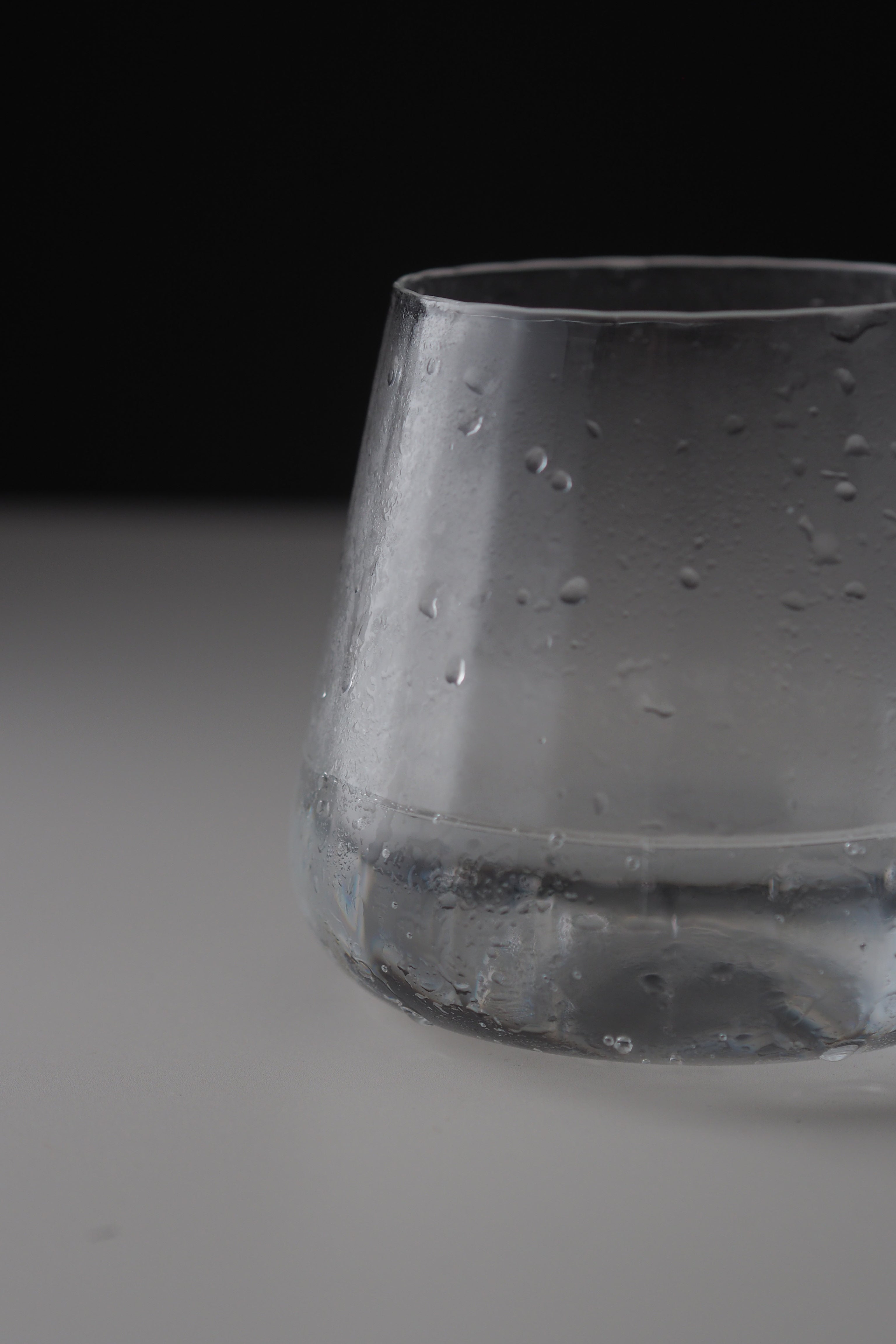 A close-up of a clear glass tumbler with condensation droplets, set against a dark black background and a light gray surface.