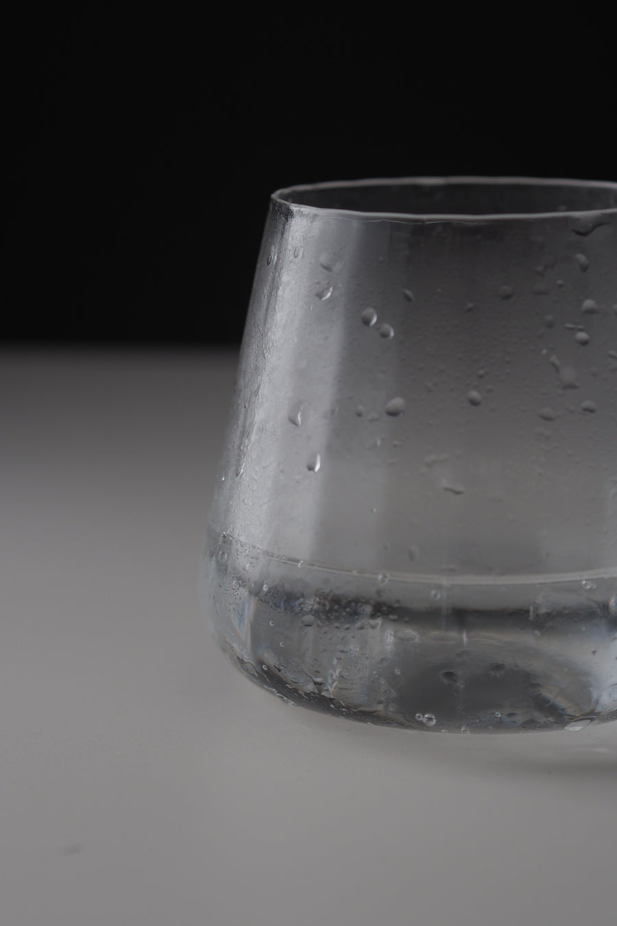 A close-up of a clear glass tumbler with condensation droplets, set against a dark black background and a light gray surface.