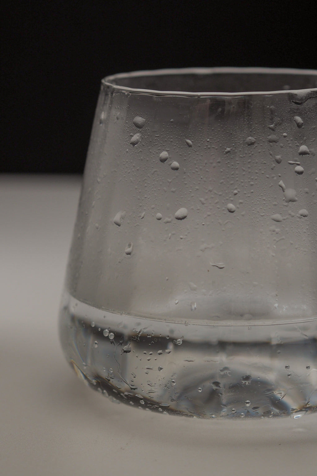A close-up of a clear glass tumbler with condensation droplets on the outside, set against a dark background and light surface.