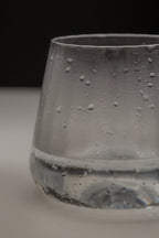 A close-up of a clear glass tumbler with condensation droplets on the outside, set against a dark background and light surface.