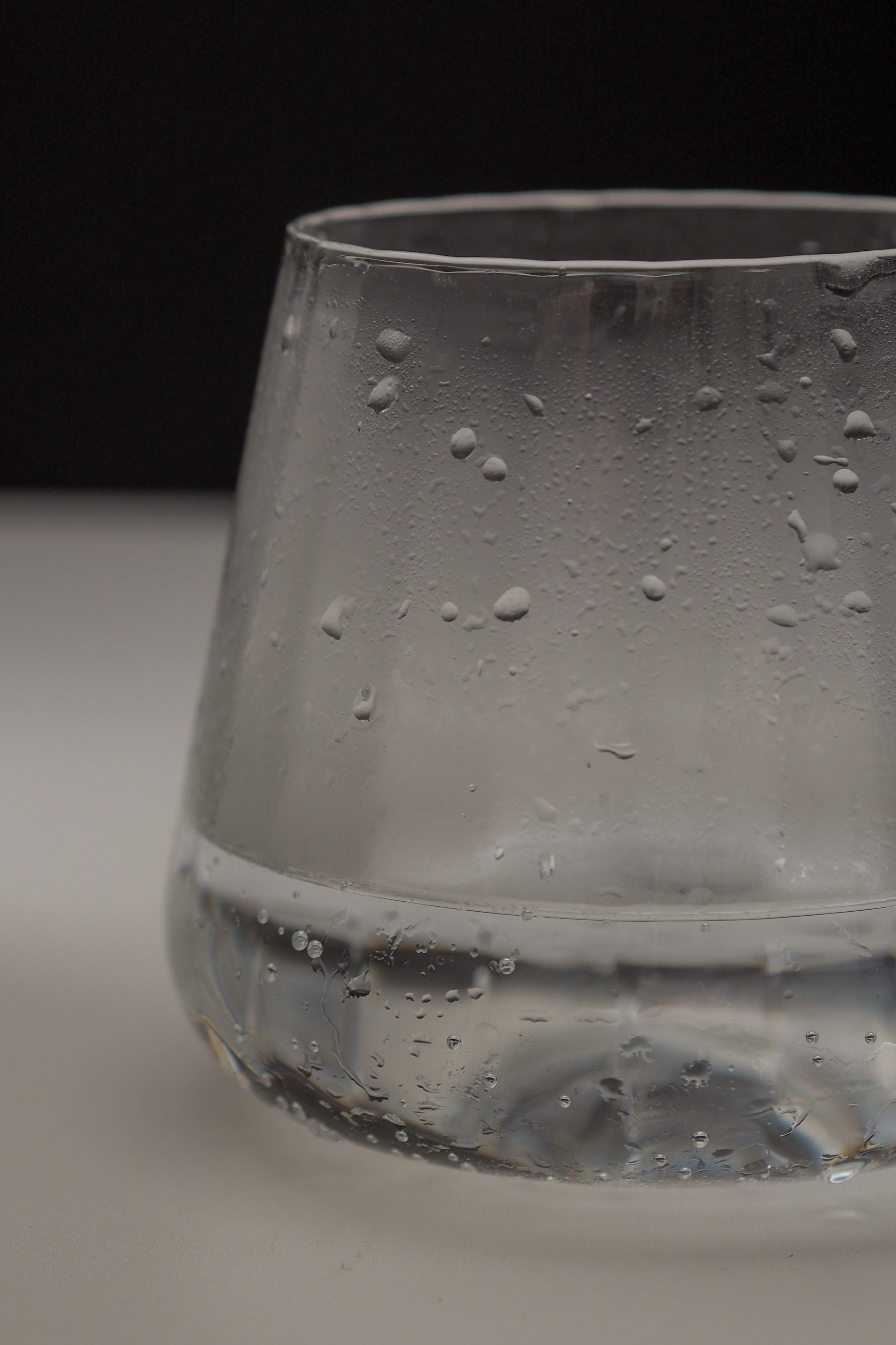 A close-up of a clear glass tumbler with condensation droplets on the outside, set against a dark background and light surface.