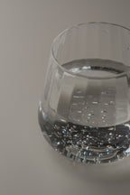 A close-up of a clear glass tumbler containing sparkling water, photographed on a plain light gray background.