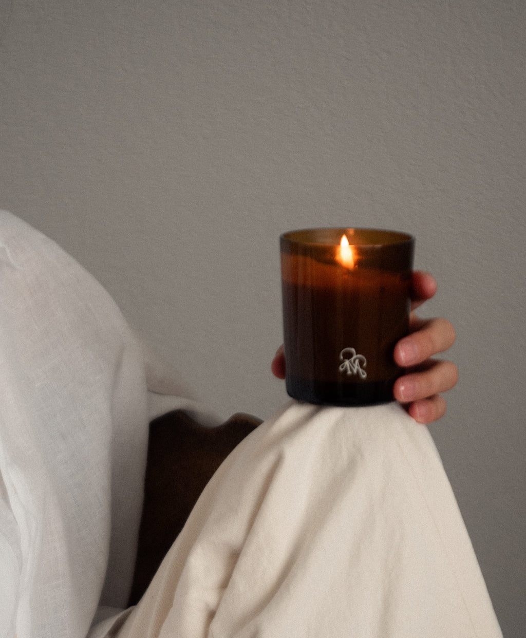 A hand holds a lit amber glass candle with a small white logo, wrapped in a cream-colored fabric against a neutral gray background.