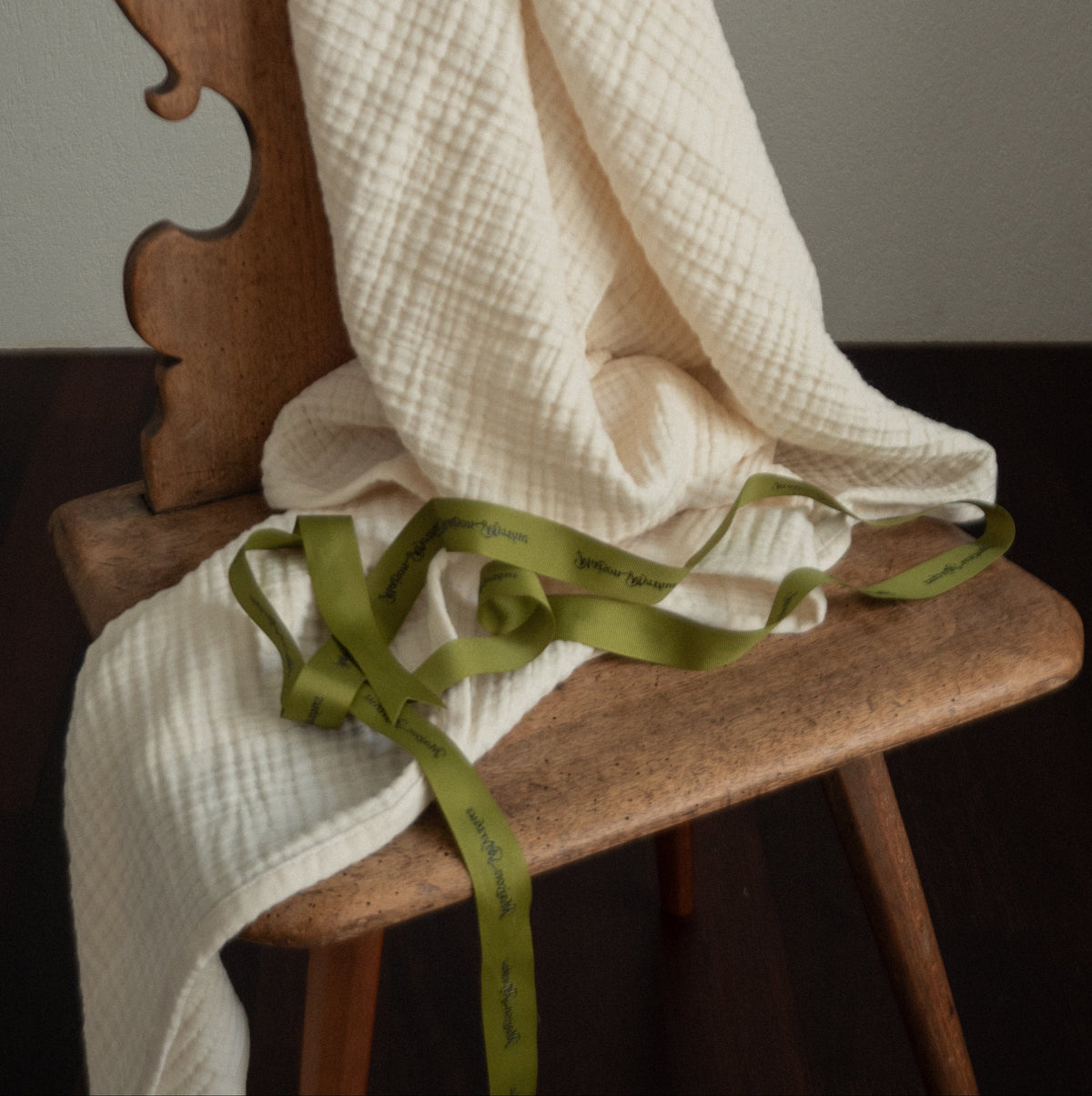 A cream-colored textured muslin blanket draped over a rustic wooden chair, tied with an olive green ribbon printed with the brand name 'Maison Myrthus'.