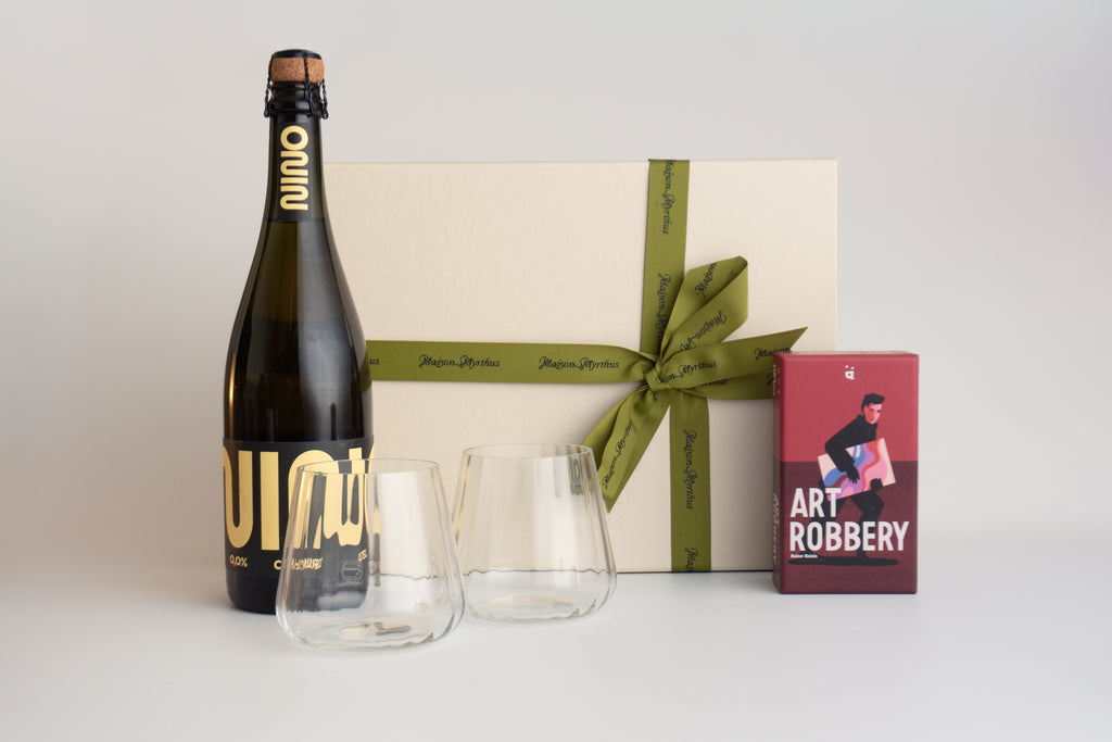 A curated gift set featuring a bottle of Quinza sparkling wine, two clear stemless wine glasses, a cream-colored gift box with an olive green ribbon, and a red 'Art Robbery' art book, arranged on a light gray background.