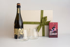 A curated gift set featuring a bottle of Quinza sparkling wine, two clear stemless wine glasses, a cream-colored gift box with an olive green ribbon, and a red 'Art Robbery' art book, arranged on a light gray background.