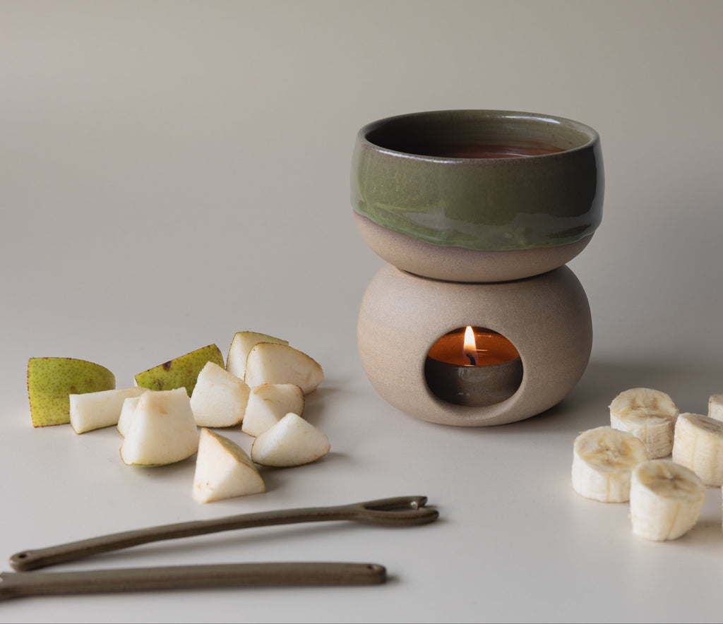 Ceramic candle burner with a candle, surrounded by fruit slices on a neutral background, for chocolate fondue