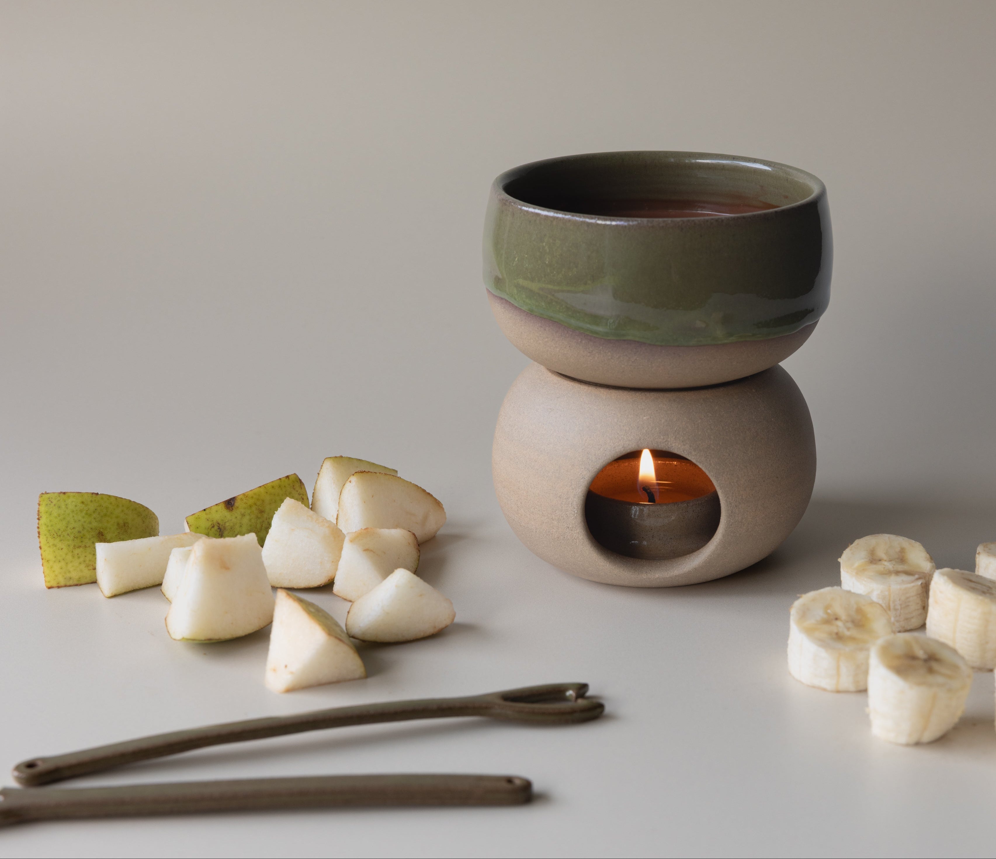 Ceramic candle burner with a candle, surrounded by fruit slices on a neutral background, for chocolate fondue