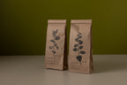Two brown paper bags with leaf designs on a green background. herbal tea made in switzerland.