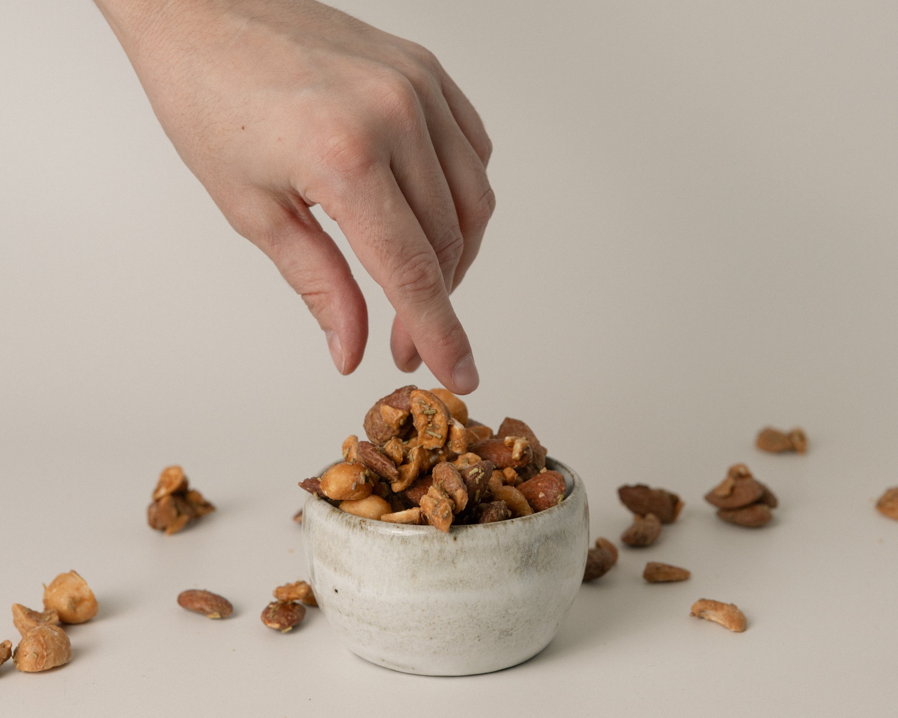 Hand reaching into a small concrete bowl filled with roasted nuts on a neutral background