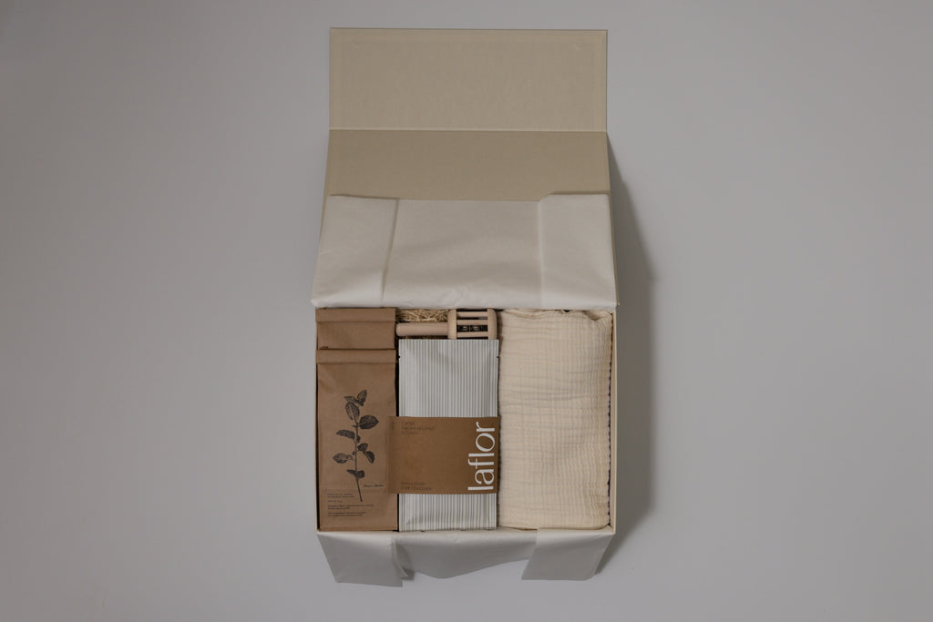 Box containing muslin blanket, laflor chocolate, herbal tea and baby rattle with a visible brand name on a plain background