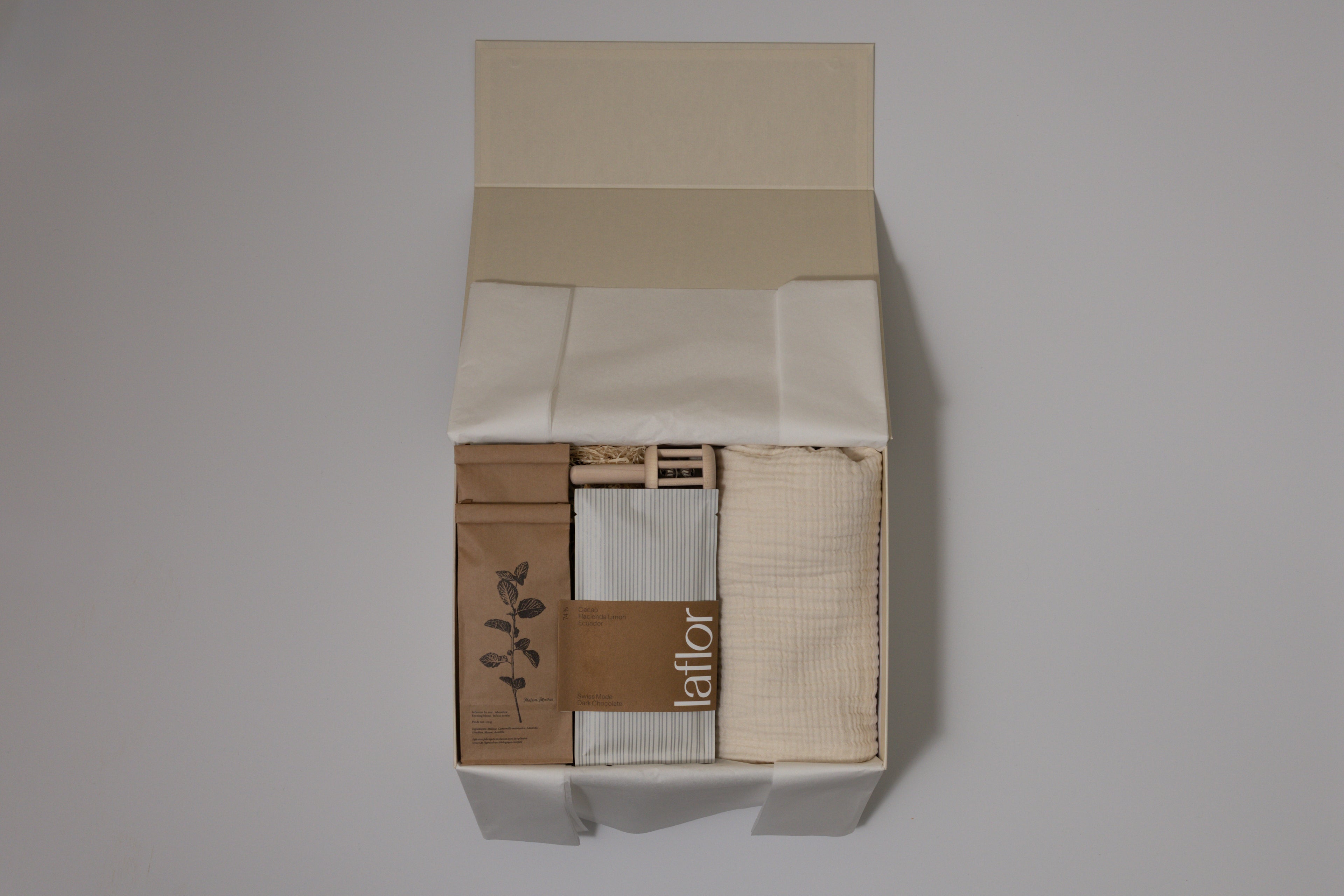 Box containing muslin blanket, laflor chocolate, herbal tea and baby rattle with a visible brand name on a plain background