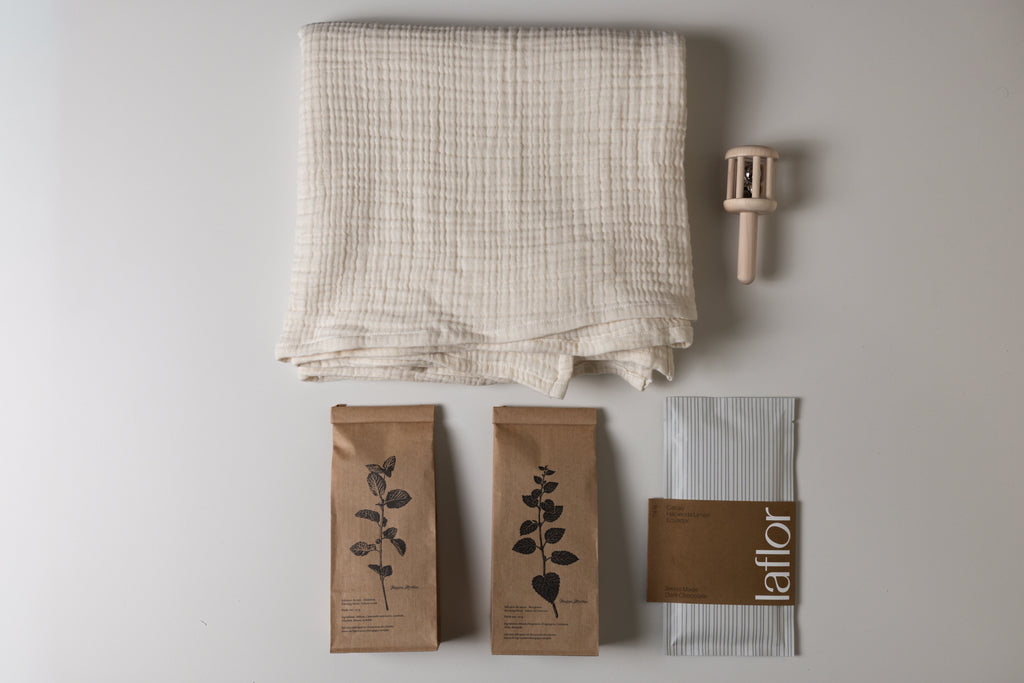 Set of items including a folded fabric, two herbal tea in kraft bags with leaf designs, and a wooden baby rattle, and laflor chocolate on a light gray background.