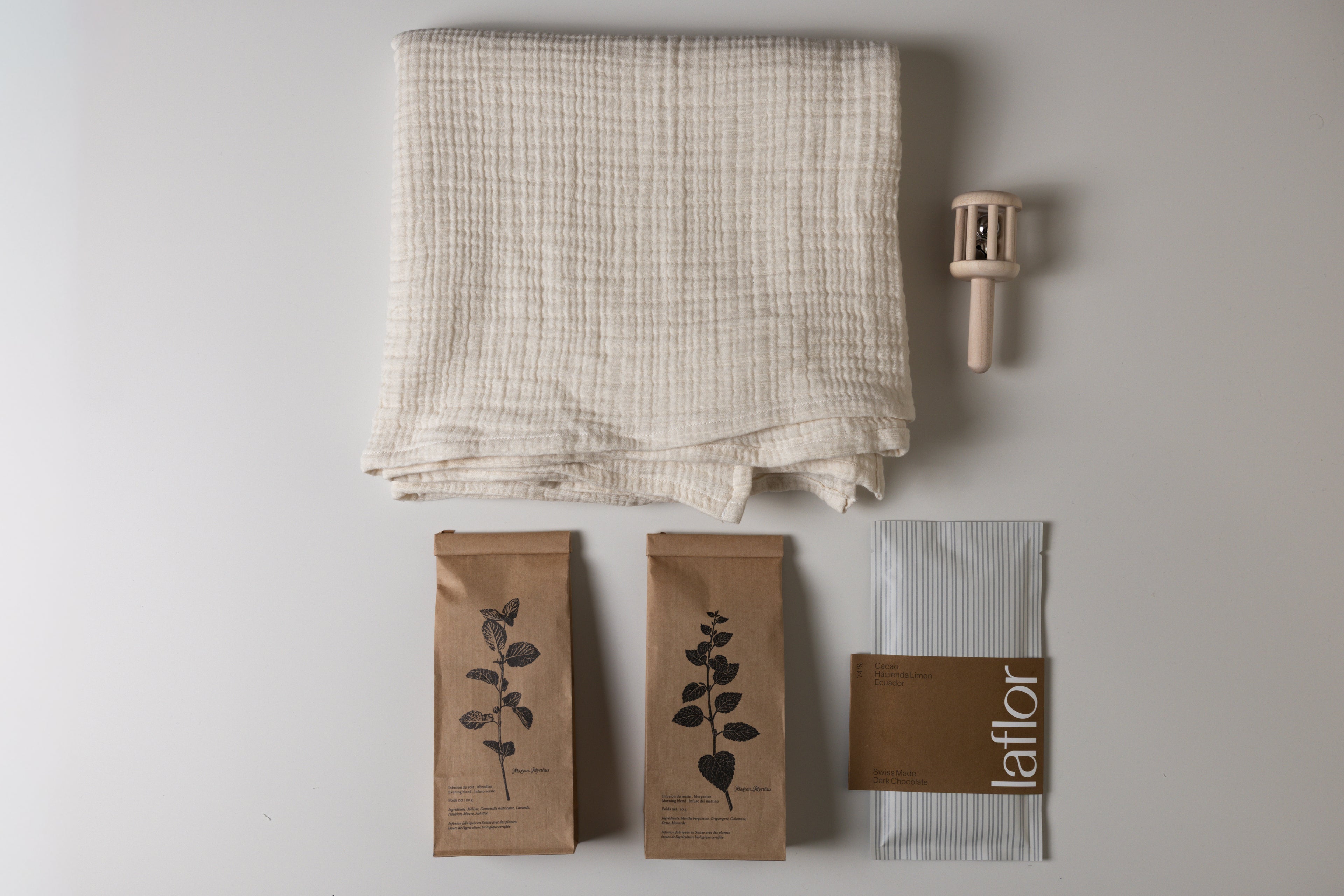 Set of items including a folded fabric, two herbal tea in kraft bags with leaf designs, and a wooden baby rattle, and laflor chocolate on a light gray background.