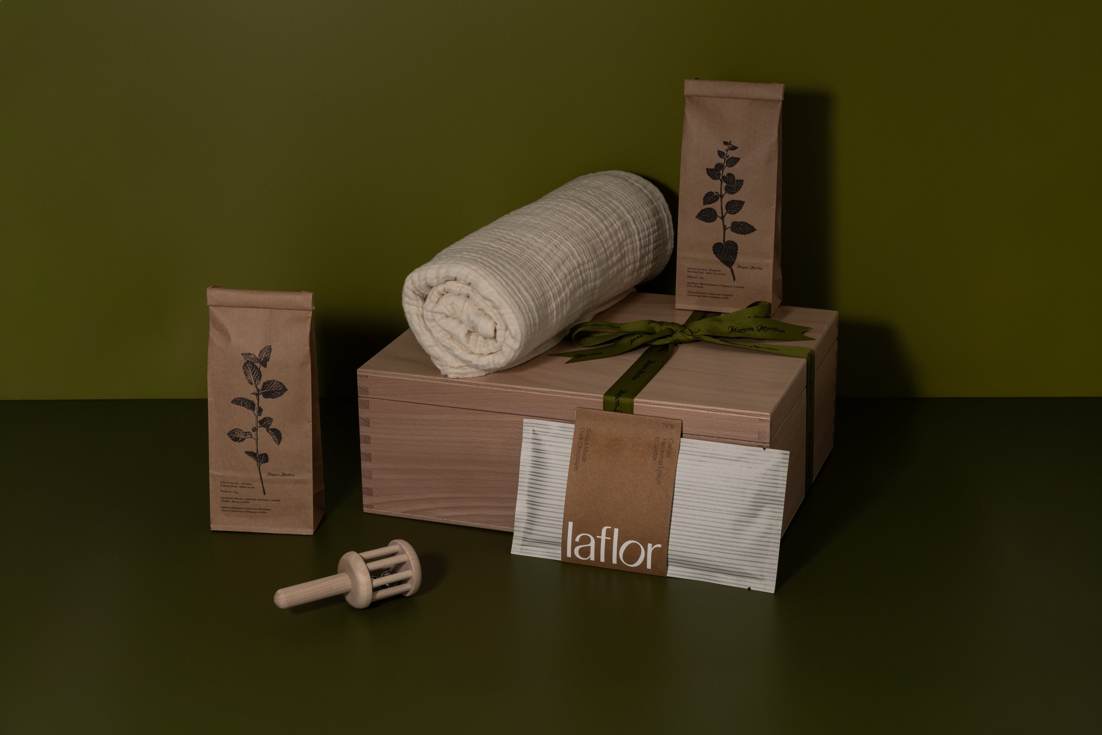 Product packaging with rolled fabric, wood boxes, and baby rattle, herbal infusion, on a dark surface against a green background