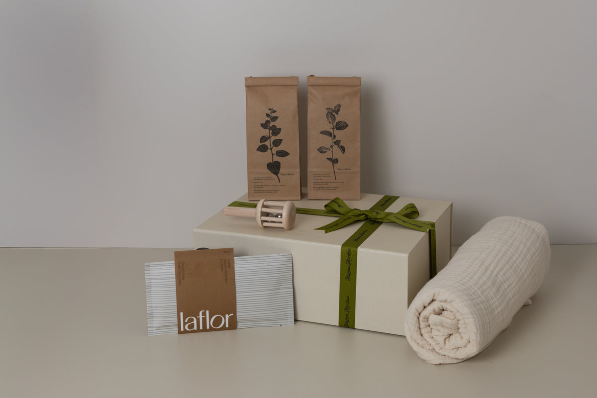 Gift box with a green ribbon, rolled baby blanket, and branded beige boxes on a neutral background, 2 tea bags, a baby rattle and laflor chocolate