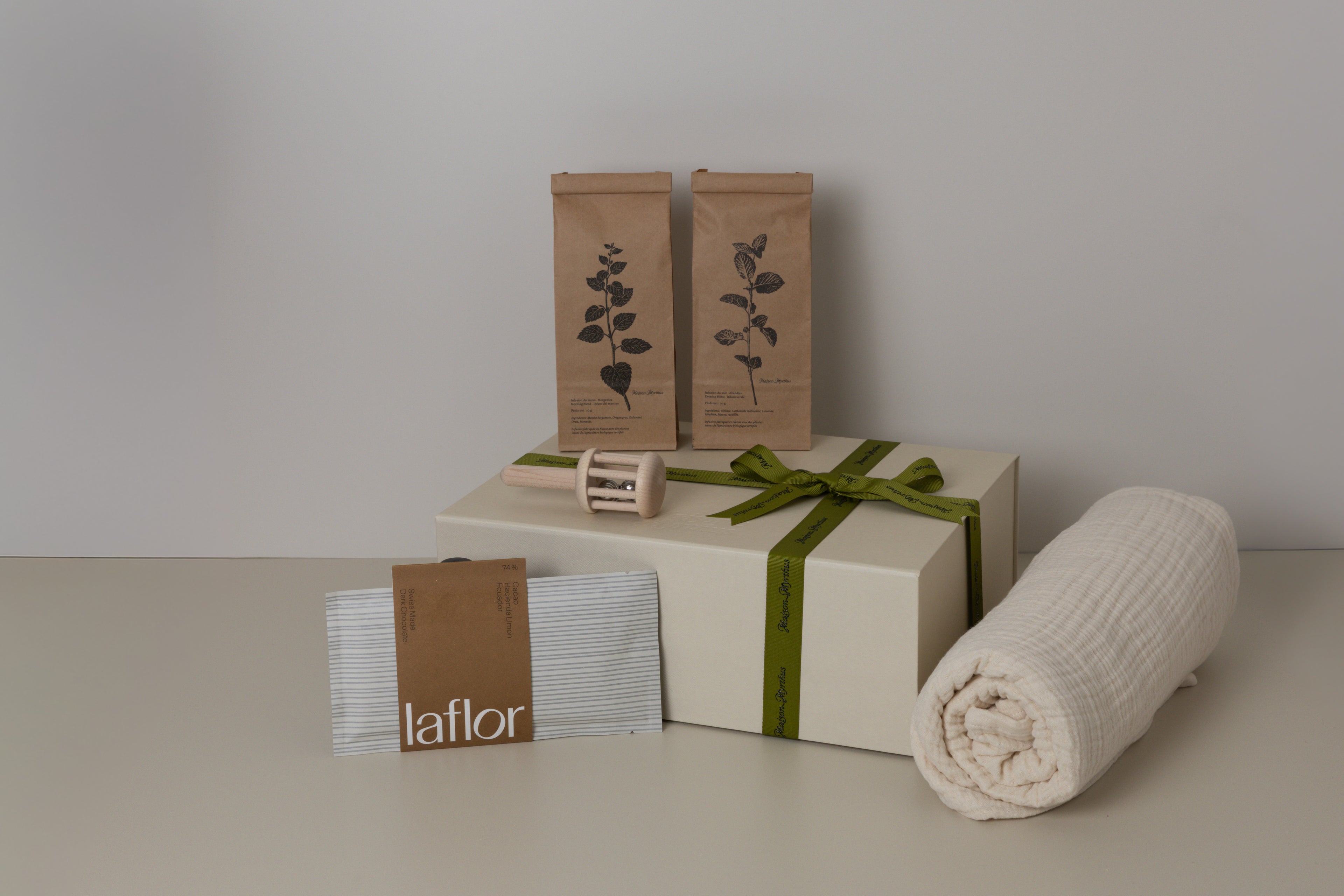 Gift box with a green ribbon, rolled baby blanket, and branded beige boxes on a neutral background, 2 tea bags, a baby rattle and laflor chocolate